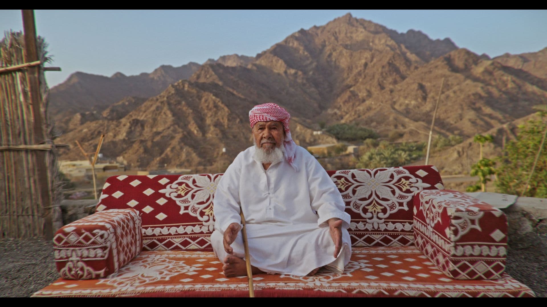 An elder seated on a red majlis before the Hajar mountains